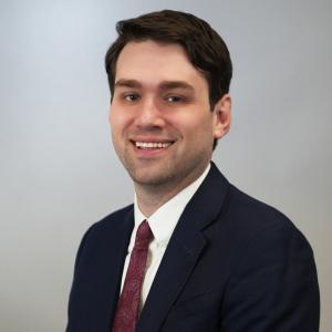 Clifford in a suit with dark red tie smiling with a white gray gradient